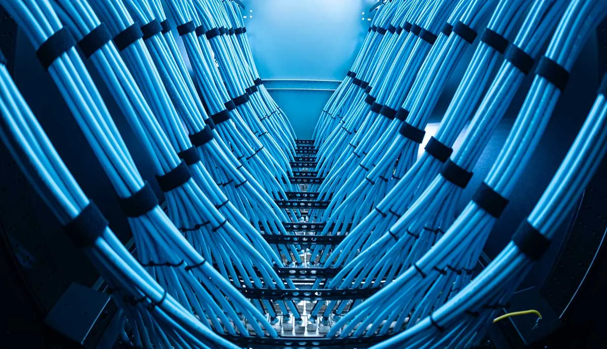 Neatly bundled blue Ethernet cables, secured with black ties, run in symmetrical rows toward network ports in a server rack, lit by cool blue overhead lights.
