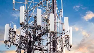 Close-up of a telecommunications tower, featuring multiple white cellular antennas and a dense network of black cables, set against a partly cloudy sky.