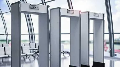 Three gray, walk-through metal detectors in an empty, modern airport terminal with large windows and rows of waiting chairs.