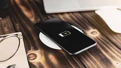 A black smartphone charging on a white wireless pad sits on a dark wooden desk, with a laptop and eyeglasses in the background.