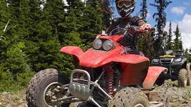 A person wearing a helmet and goggles sits on a red ATV on a dirt trail, with pine trees in the background.