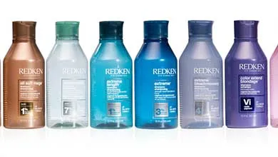 Several Redken shampoo bottles in bronze, clear, blue, and purple are lined up against a white background.