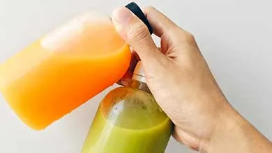 A hand clinks together an orange juice bottle and a green juice bottle against a white background.