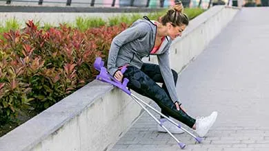 A woman in athletic clothing sits on a concrete ledge outdoors, holding her lower leg in pain. Purple crutches lie beside her on the sidewalk.