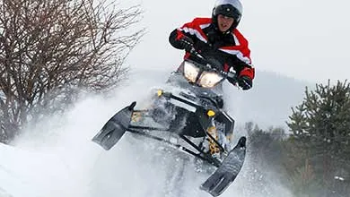 A person in a helmet and red-and-black jacket jumps a snowmobile, sending snow into the air in a snowy forest.