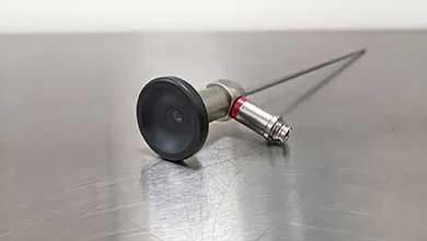 A medical endoscope with a black eyepiece and a long, thin metal shaft lies on a brushed stainless steel surface.