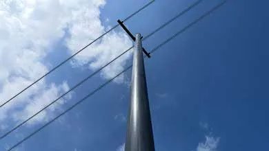 Low-angle view of a tall, dark utility pole against a bright blue sky with scattered white clouds. Three power lines run diagonally from a crossbar near the top.