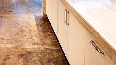 White cabinets with metallic bar handles sit below a light stone countertop, next to a polished concrete floor with tan and brown mottling.