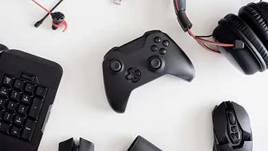 Top-down view of black gaming gear on a white surface: a central video game controller, over-ear headphones with red cables above, a partial mechanical keyboard on the left, and a gaming mouse on the right.